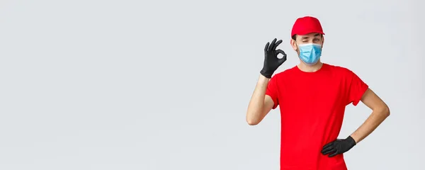 Covid-19, zelf-quarantaine, online winkelen en verzending concept. Leverancier in rood t-shirt, pet met gezichtsmasker en handschoenen, goed teken, knipoog, geen probleem. Koerier omgaan met elke overdracht — Stockfoto