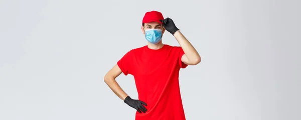 Covid-19, kwarantanna własna, zakupy online i koncepcja wysyłki. Dostawca czerwony mundur, dotykając czapki jako salutowanie klienta, pracując w wybuchu koronawirusu, nosić maskę medyczną i rękawice ochronne — Zdjęcie stockowe