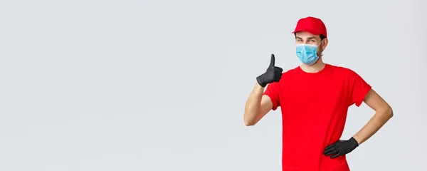 Covid-19, self-quarantine, online shopping and shipping concept. Friendly cheerful delivery guy in red uniform, gloves and face mask guarantee courier service quality, show thumb-up in like, approval — Stock Photo, Image