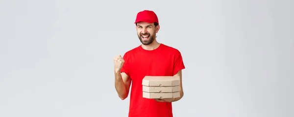 食品交付、检疫、待在家里和网上订购的概念.热情洋溢的信使身穿红色T恤，头戴红帽子，带着比萨的订单上门，手握水泵自信，赢得了庆祝胜利 — 图库照片