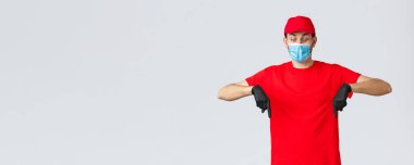 Covid-19, kendini karantina, online alışveriş ve nakliye konsepti. Üniformalı, kırmızı şapkalı ve tişörtlü ürkmüş ve şok olmuş bir kurye. Güvenlik önlemi takıyor. Tıbbi maske ve eldiven takıyor.