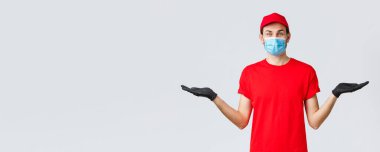 Erzak ve paket teslimatı, covid-19, karantina ve alışveriş konsepti. Kırmızı üniformalı, maskeli ve eldivenli arkadaş canlısı teslimatçı elinde iki eşya ya da afiş, tavsiye ikramiyeleri.