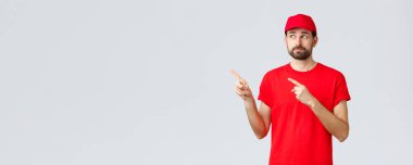 İnternetten alışveriş, karantina sırasında teslimat ve paket servis konsepti. Belirsiz ve hoşnutsuz, isteksiz kurye, kırmızı tişörtlü ve şapkalı çalışan, sol üst köşedeki parmaklarını göstererek hayal kırıklığına uğramış sırıtış.