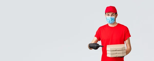 Food delivery, application, online grocery, contactless shopping and covid-19 concept. Cheerful courier in red uniform, face mask and gloves pointing finger at pizza boxes, deliver to client house — Stock Photo, Image