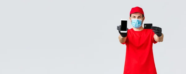 Απεριόριστη παράδοση, πληρωμή και online αγορές κατά τη διάρκεια covid-19, αυτο-καραντίνα. Φιλικός κούριερ με κόκκινη στολή, μάσκα προσώπου και γάντια, εμφάνιση οθόνης smartphone και πιστωτικής κάρτας, παραγγελία internet — Φωτογραφία Αρχείου