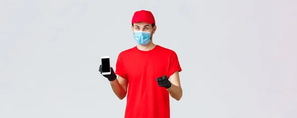 Consegna senza contatto, pagamento e shopping online durante la covid-19, auto-quarantena. Corriere sorridente in uniforme rossa, guanti e maschera viso, mostrando le informazioni sull'ordine sullo schermo del telefono, puntando la fotocamera — Foto Stock