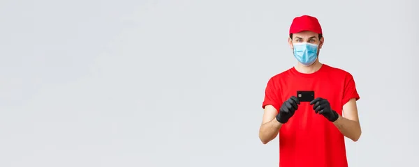 Contactless delivery, payment and online shopping during covid-19, self-quarantine. Cheerful courier in red uniform cap and t-shirt, wear medical face mask, gloves showing credit card, order food — Stock Photo, Image