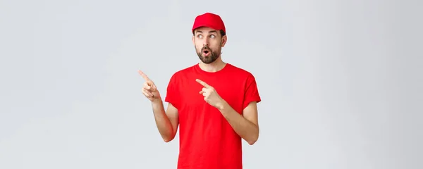 オンラインショッピング、隔離中の配達とテイクアウトの概念。帽子、従業員の制服、口を開けてショックを受け、左を見つめ、指を指している赤いTシャツの中で感動し、疑問に思った宅配便 — ストック写真