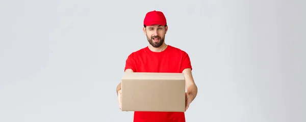 Entrega de pedidos, compras en línea y concepto de envío de paquetes. Cheeky guapo mensajero barbudo en uniforme rojo, entregando paquete de caja al cliente. Empleado guiñar a usted y dar orden paquete —  Fotos de Stock