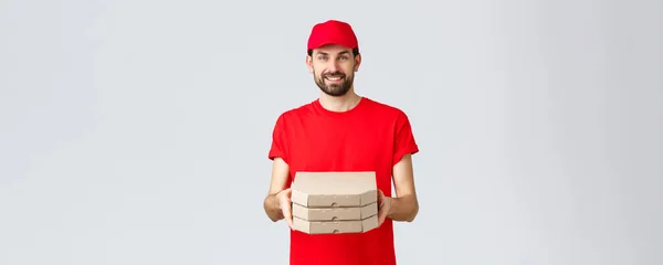 Dostawa żywności, kwarantanna, zostań w domu i zamów koncepcję online. Uśmiechnięty brodaty kurier w czerwonym mundurze czapka i t-shirt, wręczanie klientom pudełka z pizzą, dostarczyć zamówienie, szare tło — Zdjęcie stockowe