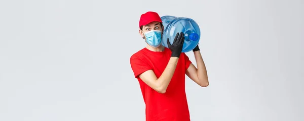 Takeaway, food and groceries delivery, covid-19 contactless orders concept. Surprised courier in red uniform cap, gloves and face mask, staring shocked as bring bottled water to office or home — Stock Photo, Image