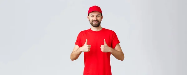 Nätshopping, leverans under karantän- och hämtningskoncept. Glada kurir i röd uniform mössa och t-shirt, rekommenderar att göra beställningar, tummen upp i godkännande, grå bakgrund — Stockfoto
