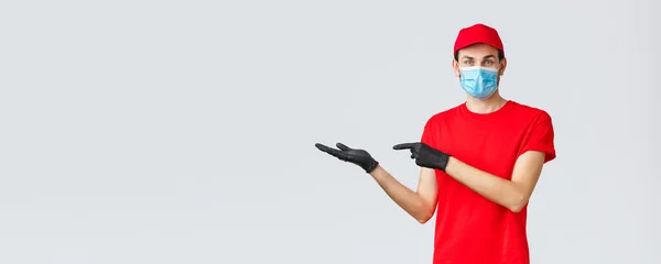 Groceries and packages delivery, covid-19, quarantine and shopping concept. Friendly courier in red uniform, face mask and gloves, introduce promo on banner, pointing left, showing advertisement — Stock Photo, Image