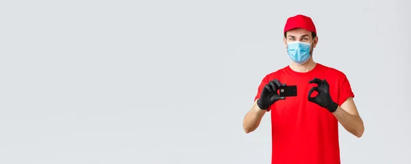 Contactless delivery, payment and online shopping during covid-19, self-quarantine. Friendly courier in red uniform, cap and t-shirt, wear face mask and gloves, thumb-up recommend credit card paying — Stock Photo, Image