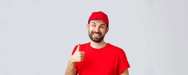 Online shopping, delivery during quarantine and takeaway concept. Smiling handsome bearded delivery guy or courier in red uniform cap, show supportive thumb-up, guarantee safe order transfer — Stock Photo, Image