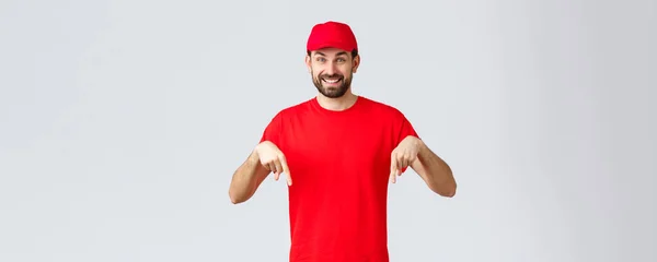 オンラインショッピング、隔離中の配達とテイクアウトの概念。赤い制服キャップとTシャツの陽気なフレンドリーな宅配便,指を下に指して,クライアントに通知,広告をお勧めします — ストック写真