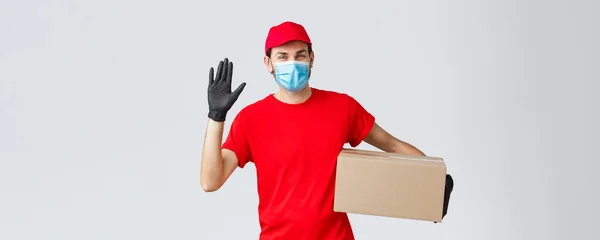 Consegna pacchi e pacchi, consegna covid-19 in quarantena, ordini di trasferimento. Gentile corriere in uniforme rossa, maschera viso con guanti protettivi, consegna scatola ordini al cliente, mano agitante in ciao — Foto Stock