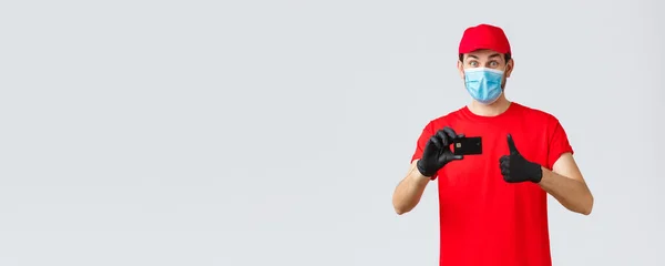 Contactless delivery, payment and online shopping during covid-19, self-quarantine. Excited happy courier in red t-shirt and cap uniform, face mask with gloves, recommend credit card order paying — Stock Photo, Image