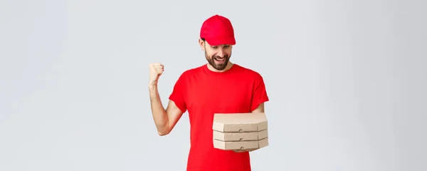 食料の配達、隔離、家にいてオンラインの概念を注文しなさい。喜んでピザの注文を見て灰色の背景に赤いTシャツとキャップの幸せな興奮したひげそりの宅配業者 — ストック写真