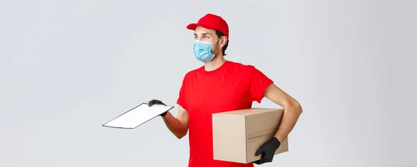 Packages and parcels delivery, covid-19 delivery, transfer orders. Profile of friendly courier in red uniform in face mask and gloves, bring order, hold box and give form for client to sign — Stock Photo, Image