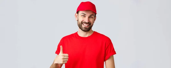 Online winkelen, levering tijdens quarantaine en afhaalconcept. Vriendelijke, vrolijke koerier in rood mutsje en t-shirt uniform, aan te moedigen maken internet bestellingen, thumb-up in goedkeuring of aanbeveling — Stockfoto