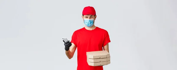Food delivery, application, online contactless shopping and covid-19 concept. Courier in red uniform, face mask and gloves, winking to client, inform bonuses, special discounts on pizza, hold phone — Stock Photo, Image