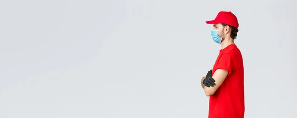 Covid-19, αυτο-καραντίνα, online αγορές και ναυτιλία έννοια. Προφίλ f καθορισμένο courier σε κόκκινο t-shirt και καπέλο, στολή του φορέα, σταυρό στο στήθος χέρια, μετατόπιση εργασίας, give-out παραγγελίες πελατών — Φωτογραφία Αρχείου