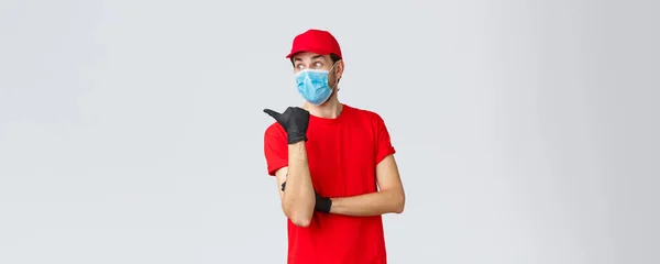 Covid-19, self-quarantine, online shopping and shipping concept. Surprised courier in red uniform, face mask and gloves, pointing thumb and look left impressed, curiously reading promo, show way — Stock Photo, Image