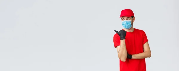 Covid-19, self-quarantena, shopping online e concetto di spedizione. Consegna ragazzo in uniforme rossa, guanti e maschera viso, puntando a sinistra e sorridente, consiglia di utilizzare il servizio di corriere client — Foto Stock