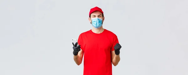 Consegna senza contatto, pagamento e shopping online durante la covid-19, auto-quarantena. Gioia, felice corriere in uniforme rossa, guanti e maschera, sorridente, leggere buone notizie in applicazione, cellulare — Foto Stock