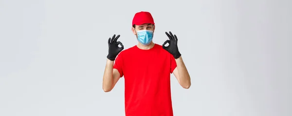Covid-19, αυτο-καραντίνα, online αγορές και ναυτιλία έννοια. Όμορφος ντελιβεράς με κόκκινο καπέλο, t-shirt, φοράει προστατευτική μάσκα προσώπου και γάντια από το coronavirus, δείχνουν εντάξει, συμφωνούν και καλή χειρονομία — Φωτογραφία Αρχείου