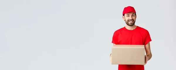 Bestil levering, online shopping og pakke shipping koncept. Venlig smilende kurer i rød ensartet hætte og t-shirt, uddele pakker til kunder. Medarbejder medbringer pakkeæske, grå baggrund - Stock-foto