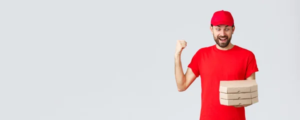 Food delivery, quarantine, stay home and order online concept. Excited and happy courier in red t-shirt and cap, rejoicing holding tasty pizzas, look at boxes and fist pump, grey background — Stock Photo, Image