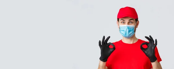 Contactless delivering, covid-19 and shopping concept. Cheerful, pleased courier in red uniform, cap and medical mask with gloves show okay, approval or guarantee gesture, recommend service — Stock Photo, Image
