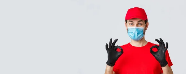 Contactloze levering, covid-19 en shopping concept. Vrolijke, tevreden koerier in rood uniform, pet en medisch masker met handschoenen te tonen oke, goedkeuring of garantie gebaar, raden service — Stockfoto