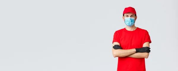 Covid-19, self-quarantine, online shopping and shipping concept. Confident smiling delivery man in red cap, t-shirt, wearing protective medical mask and rubber gloves while making courier order — Stock Photo, Image