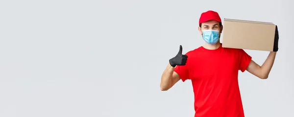 Packages and parcels delivery, covid-19 quarantine delivery, transfer orders. Cheerful courier in red uniform, gloves and face mask, thumb-up, no problem deliver order box to customers — Stock Photo, Image