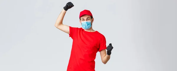 Groceries and packages delivery, covid-19, quarantine and shopping concept. Cheerful courier red uniform, gloves and face mask, raising hands up, chanting rejoicing over good news, fight coronavirus — Stock Photo, Image