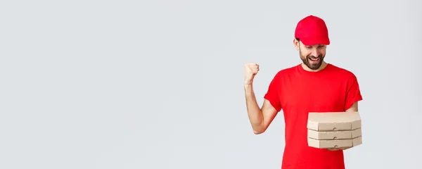 Food delivery, quarantine, stay home and order online concept. Happy, excited bearded courier in red t-shirt and cap celebrating, rejoicing, looking at pizza orders, grey background — Stock Photo, Image