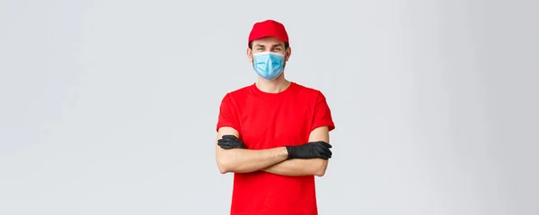 Covid-19, kwarantanna własna, zakupy online i koncepcja wysyłki. Pewny siebie uśmiechnięty dostawca w czerwonej czapce, koszulce, noszący ochronną maskę medyczną i gumowe rękawiczki podczas składania zamówienia kuriera — Zdjęcie stockowe