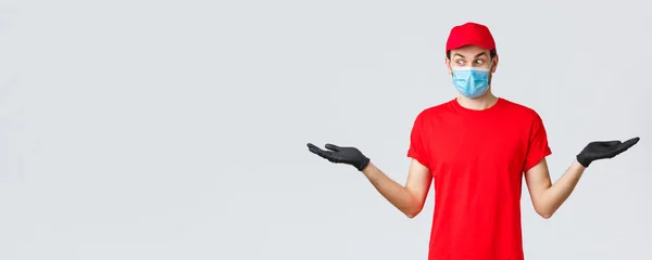 Παντοπωλεία και παράδοση πακέτων, covid-19, καραντίνα και εμπορική έννοια. Έκπληκτος κούριερ με κόκκινο καπέλο και t-shirt ομοιόμορφη, φορούν προστατευτική μάσκα προσώπου, γάντια, κρατήστε δύο στοιχεία στο πλάι — Φωτογραφία Αρχείου