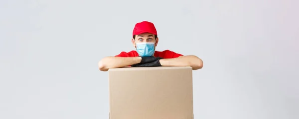 Packages and parcels delivery, covid-19 quarantine and transfer orders. Worried and confused courier in red uniform, gloves and face mask, lean on order box and look surprised camera — Stock Photo, Image