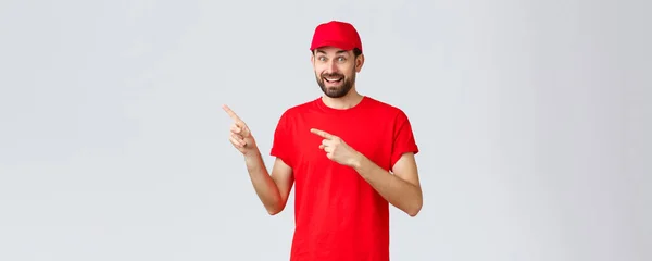オンラインショッピング、隔離中の配達とテイクアウトの概念。楽しさと興奮笑顔宅配便クライアントを見て、赤い制服キャップとTシャツの指差し指の従業員左 — ストック写真