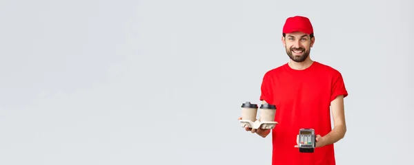 Food delivery, quarantine, stay home and order online concept. Friendly bearded courier in red uniform handing coffee delivery and POS terminal for client to pay contactless, grey background — Stock Photo, Image