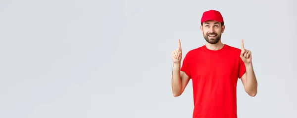 Online αγορές, παράδοση κατά τη διάρκεια καραντίνας και takeaway έννοια. Χαμογελώντας χαρούμενα κούριερ με κόκκινο μπλουζάκι, με καπέλο που δείχνει προς τα πάνω. Υπάλληλος βλέμμα ιντριγκάρει στο πανό με ειδική έκπτωση — Φωτογραφία Αρχείου