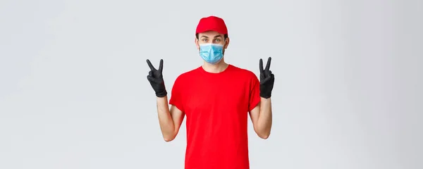 Covid-19, self-quarantine, 온라인 쇼핑 및 배송 컨셉. 빨간 유니폼을 입고 얼굴에 마스크를 쓰고 장갑을 끼고 코로나 바이러스 발병에 긍정적으로 반응하고 평화 표시를 하고 — 스톡 사진