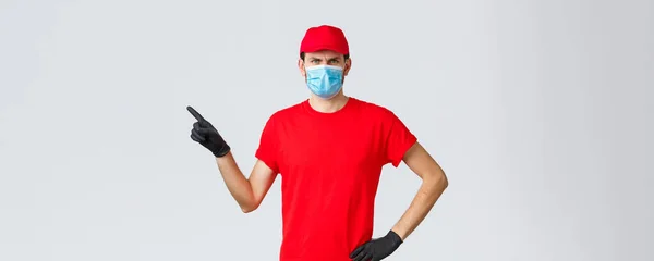 Covid-19, self-quarantine, online shopping and shipping concept. Displeased angry delivery guy scolding courier making mistake in transfer, frowning, pointing finger left, wear medical mask — Stock Photo, Image