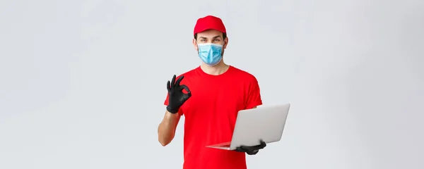 Customer support, covid-19 delivery packages, online orders processing concept. Smiling courier in face mask and gloves guarantee safety of parcel, processing order, show okay sign, hold laptop — Stock Photo, Image