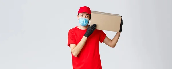 Packages and parcels delivery, covid-19 quarantine delivery, transfer orders. Curious courier in red uniform, gloves and protective face mask, deliver box to client, bring order contactless — Stock Photo, Image