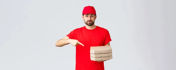 食料の配達、隔離、家にいてオンラインの概念を注文しなさい。赤い制服キャップとTシャツで自信を持ってフレンドリーな宅配便,従業員は注文ピザを持って来ます,箱で指を指して,グレーの背景 — ストック写真